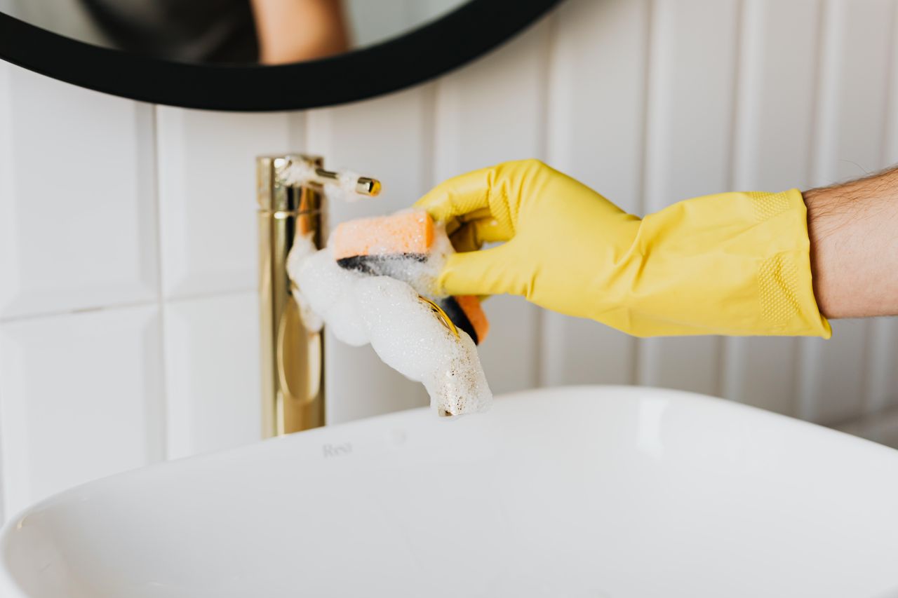 Un homme méconnaissable portant des gants en latex jaunes nettoie un robinet doré dans une salle de bains, tout en faisant le ménage à la maison avec une éponge orange et du détergent